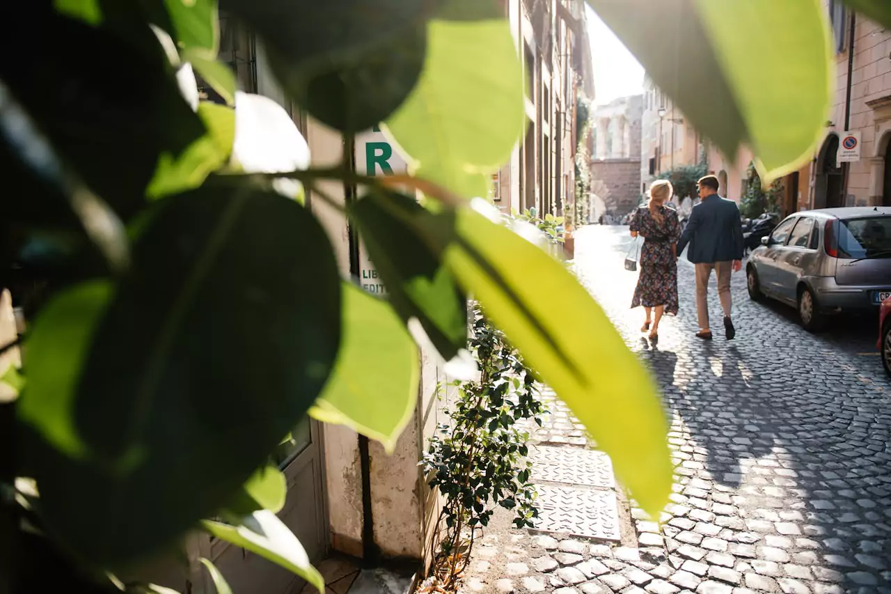 Un couple élégant marche main dans la main sur une rue pavée ensoleillée d'une ville européenne, entouré de bâtiments historiques. La scène est partiellement encadrée par de grandes feuilles vertes au premier plan, créant une atmosphère romantique et chaleureuse.