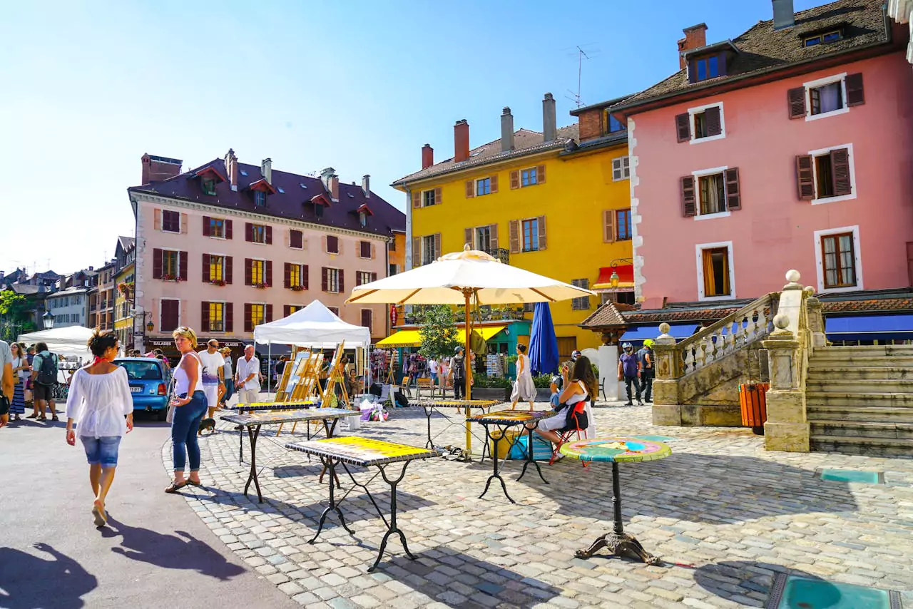 Marché animé à Annecy avec des stands d'artisanat et des bâtiments colorés en arrière-plan, sous un ciel dégagé.