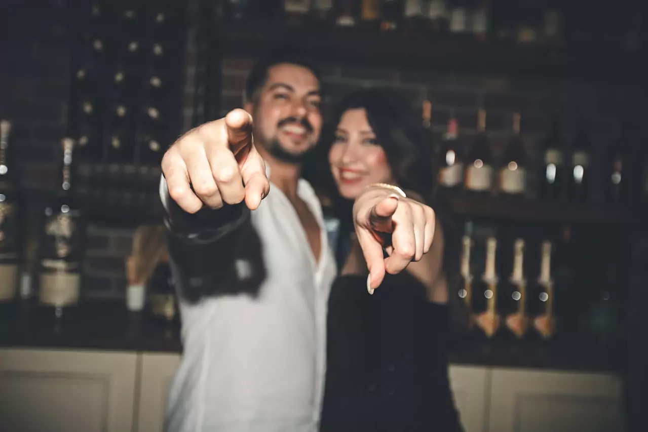 Un couple souriant et habillé élégamment dans un bar, pointant directement vers l’appareil photo avec une attitude joyeuse et complice. L'arrière-plan est flou avec des bouteilles d'alcool sur des étagères.