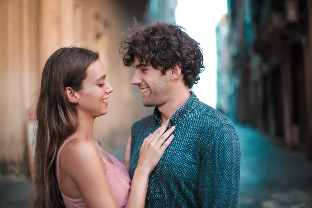 Un homme et une femme se regardent avec complicité dans une rue ancienne, échangeant un sourire tendre. L’homme aux cheveux bouclés semble heureux et captivé par la femme qui pose délicatement sa main sur sa chemise, créant une atmosphère romantique et intime.
