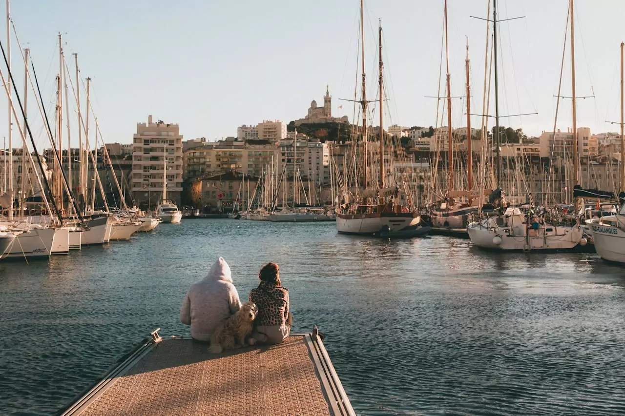 Un couple assis sur un ponton, face au Vieux-Port de Marseille au coucher du soleil, avec des voiliers amarrés et la basilique Notre-Dame de la Garde en arrière-plan. Une scène intime et paisible, idéale pour une escapade romantique.
