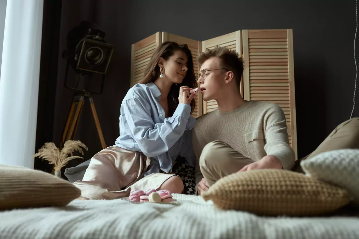 Couple complice dans une chambre coquine, assis sur un lit entouré de coussins, dans une ambiance romantique et intime
